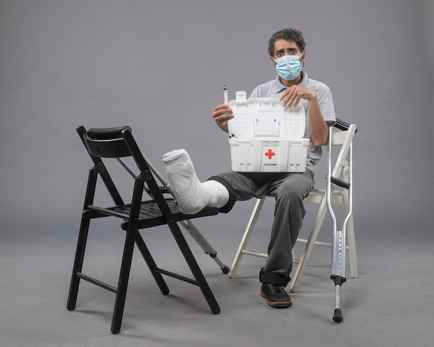 Vue de face jeune homme avec masque assis avec pied cassé et vérifiant la trousse de premiers soins sur le mur gris torsion mâle cassé la jambe du pied douloureux