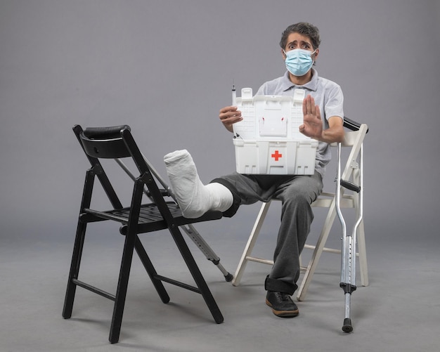 Vue de face jeune homme avec masque assis avec pied cassé et vérifiant la trousse de premiers soins sur le mur gris torsion mâle cassé la jambe du pied douloureux