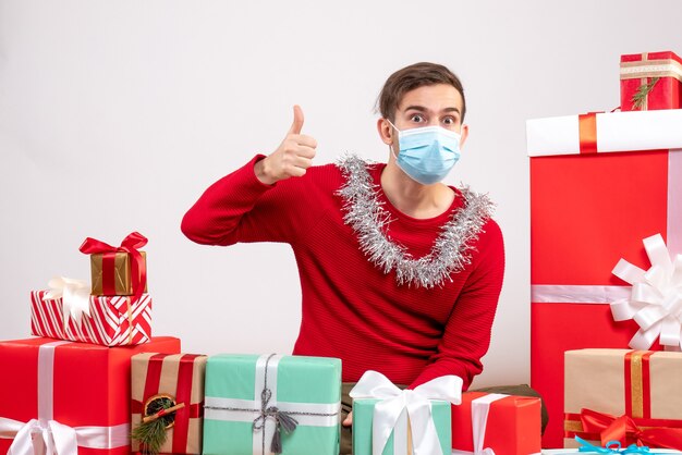 Vue de face jeune homme avec masque faisant le pouce vers le haut signe assis autour de cadeaux de Noël