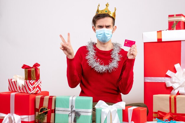 Vue de face jeune homme avec masque faisant signe de la victoire assis sur des cadeaux de Noël au sol