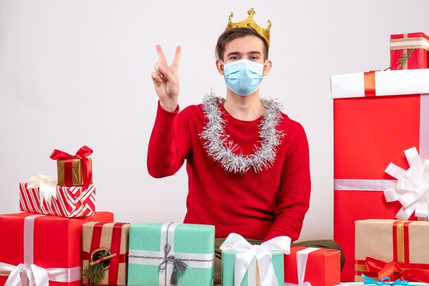 Vue de face jeune homme avec masque faisant signe de la victoire assis sur le sol autour de cadeaux de Noël