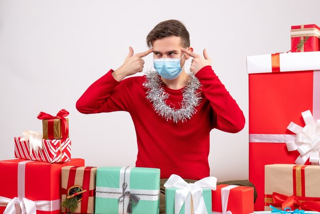 Vue de face jeune homme avec masque mettant les doigts à son temple assis autour de cadeaux de Noël