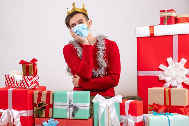 Vue de face jeune homme avec masque mettant la main sur sa joue assis sur des cadeaux de Noël au sol