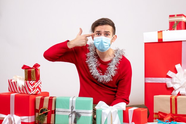 Vue de face jeune homme avec masque mettant le pistolet à doigt à son temple assis autour de cadeaux de Noël