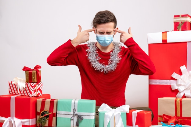 Vue de face jeune homme avec masque mettant des pistolets à doigt à son temple assis autour de cadeaux de Noël