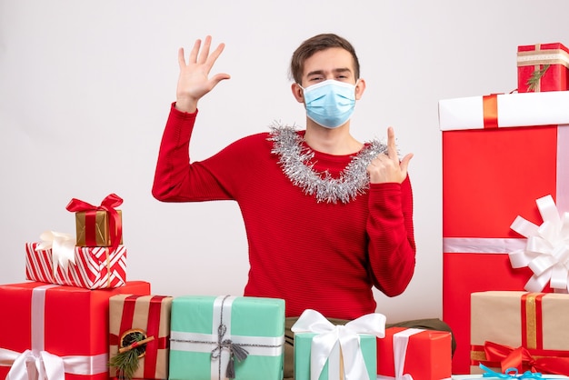 Vue de face jeune homme avec masque mettant sa main assis autour de cadeaux de Noël