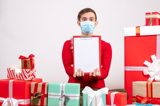 Vue de face jeune homme avec masque montrant le presse-papiers assis sur des cadeaux de Noël au sol