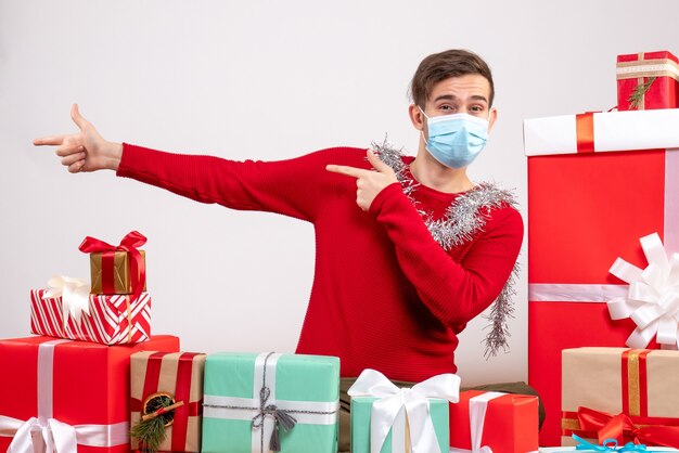 Vue de face jeune homme avec masque montrant quelque chose assis autour de cadeaux de Noël