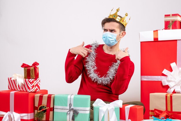 Vue de face jeune homme avec masque pointant sur lui-même assis sur des cadeaux de Noël au sol