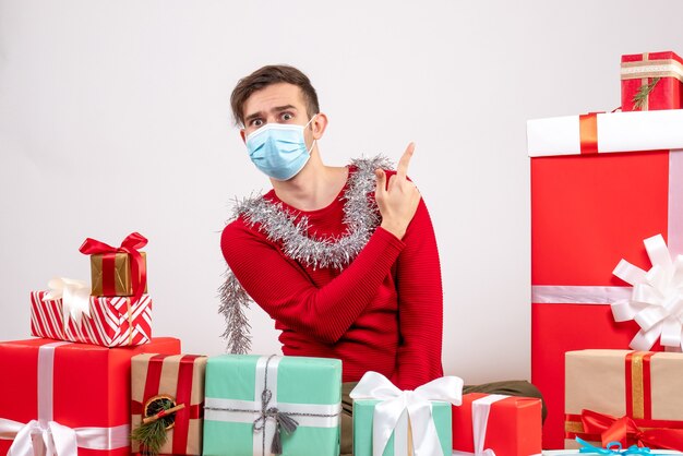 Vue de face jeune homme avec masque pointant vers l'arrière assis autour de cadeaux de Noël