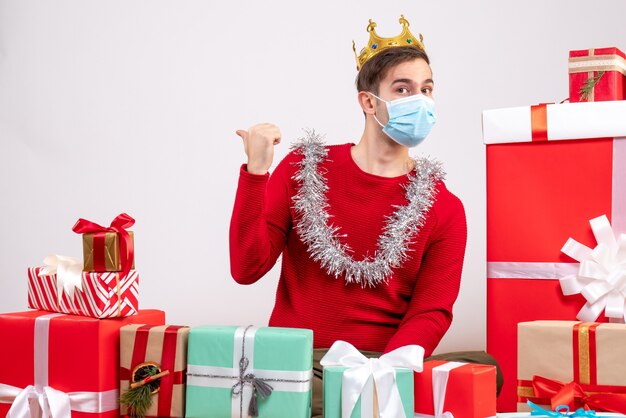 Vue de face jeune homme avec masque pointant vers l'arrière assis autour de cadeaux de Noël