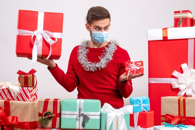 Vue de face jeune homme avec masque regardant cadeau assis autour de cadeaux de Noël