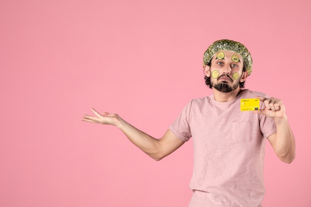 Vue de face jeune homme avec masque sur son visage tenant une carte bancaire sur fond rose