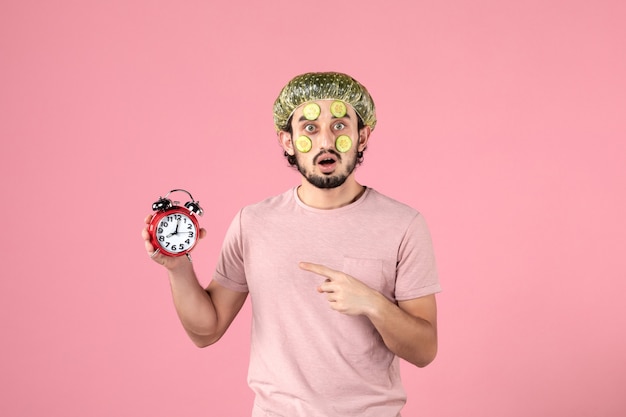 Vue de face jeune homme avec masque sur son visage tenant une horloge sur fond rose