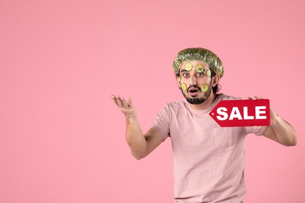 Vue de face jeune homme avec masque sur son visage tenant la plaque signalétique de vente sur fond rose