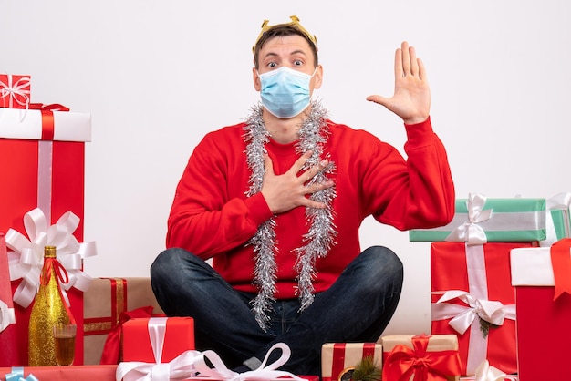 Vue de face d'un jeune homme en masque stérile assis autour de cadeaux de Noël sur un mur blanc