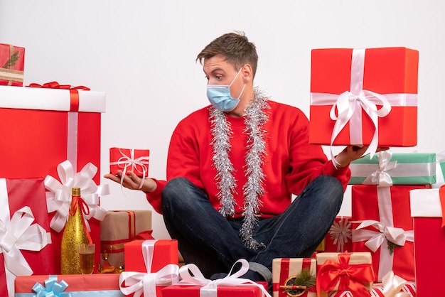 Vue de face d'un jeune homme en masque stérile assis autour de cadeaux de Noël sur un mur blanc