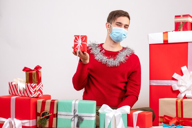 Vue de face jeune homme avec masque tenant un cadeau assis autour de cadeaux de Noël