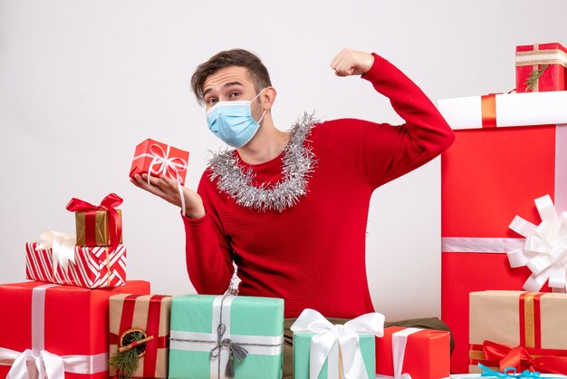 Vue de face jeune homme avec masque tenant un cadeau de Noël assis autour de cadeaux de Noël