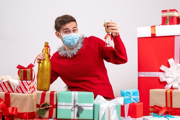 Vue de face jeune homme avec masque tenant du champagne assis autour de cadeaux de Noël