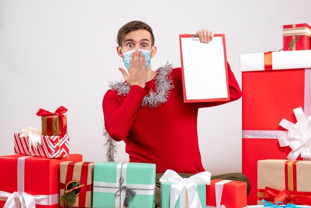 Vue de face jeune homme avec masque tenant le presse-papiers assis autour de cadeaux de Noël