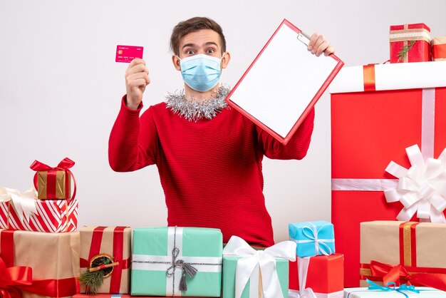 Vue de face jeune homme avec masque tenant le presse-papiers et la carte assis autour de cadeaux de Noël