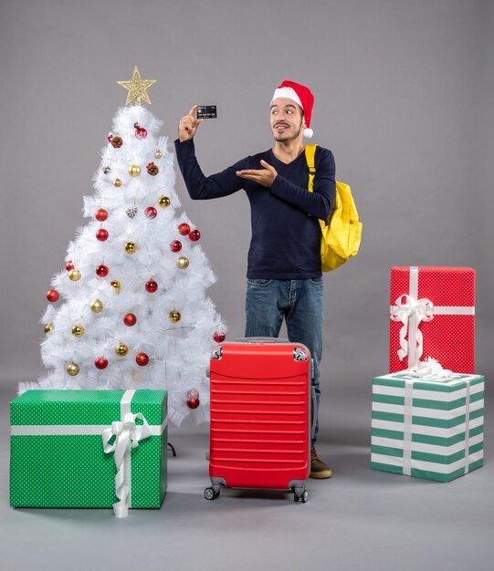 Vue de face jeune homme montrant la carte debout près de l'arbre de Noël et présente sur gris isolé