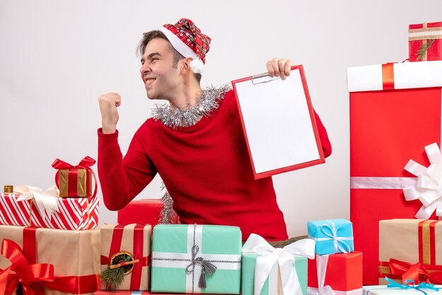 Vue de face jeune homme montrant le geste gagnant assis autour de cadeaux de Noël