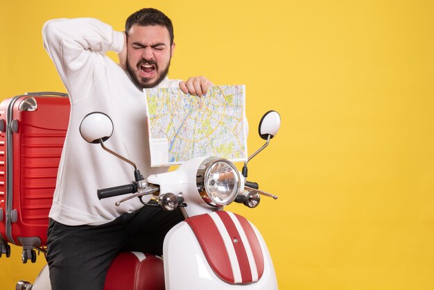 Vue de face d'un jeune homme nerveux et troublé assis sur une moto avec une valise dessus tenant une carte souffrant de douleurs à l'oreille sur fond jaune isolé