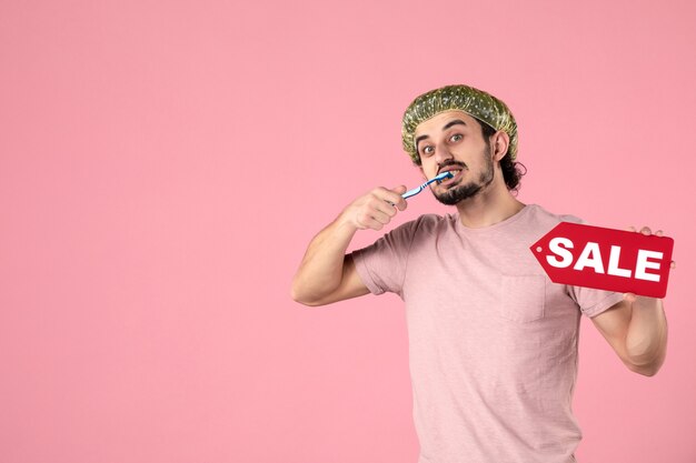 Vue de face jeune homme nettoyant ses dents et tenant la plaque signalétique de vente sur fond rose