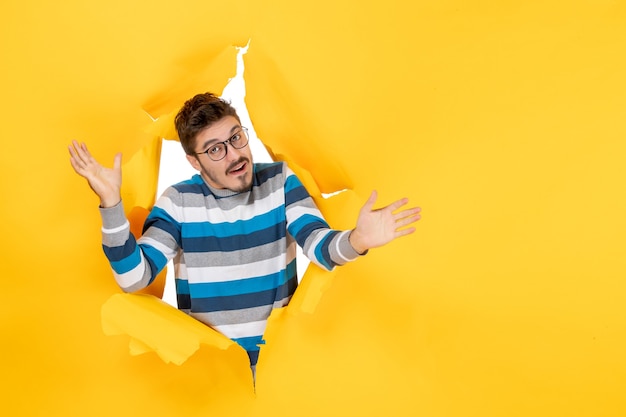 Vue de face jeune homme ouvrant les mains en regardant à travers le mur jaune en papier déchiré