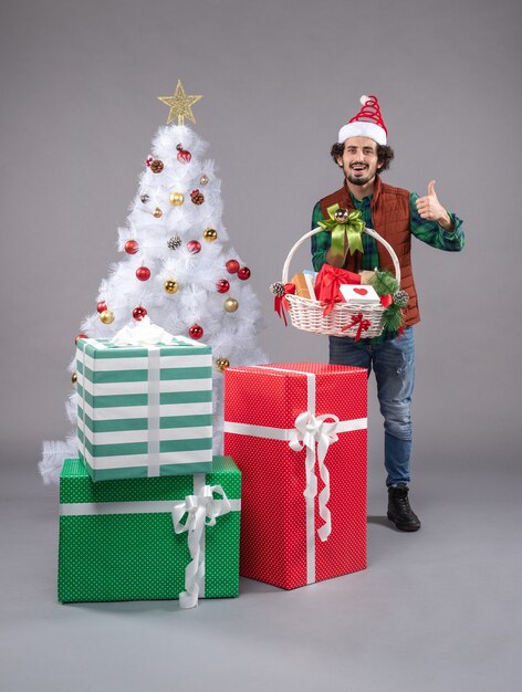 Vue de face jeune homme avec panier autour des cadeaux sur le bureau gris cadeau de Noël du nouvel an