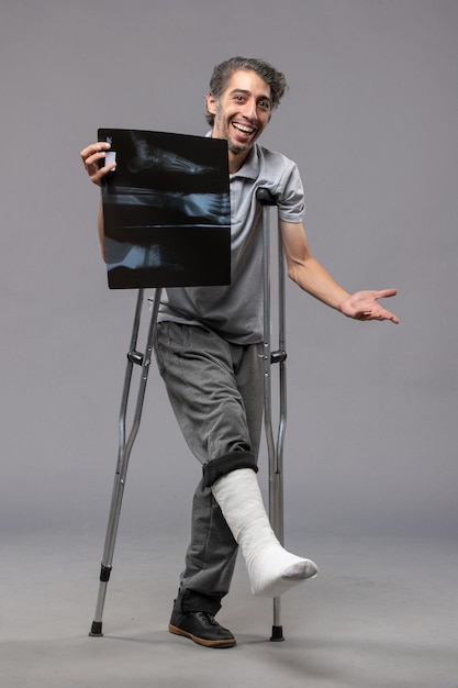 Photo gratuite vue de face jeune homme avec pied cassé à l'aide de béquilles et tenant sa radiographie sur un bureau gris désactiver la torsion du pied de douleur d'accident cassé