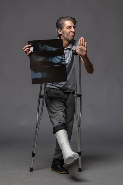 Vue de face jeune homme avec pied cassé à l'aide de béquilles et tenant sa radiographie sur un mur gris foncé accident de pied torsion douleur cassée