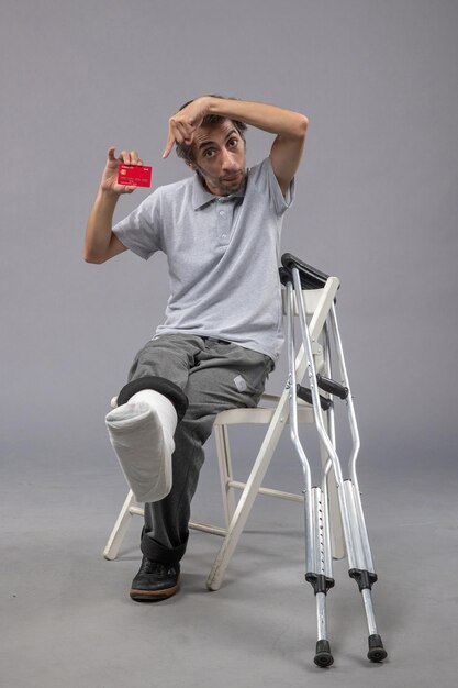 Photo gratuite vue de face jeune homme avec pied cassé et bandage tenant une carte bancaire sur un bureau gris accident torsion jambe pied douleur humaine mâle