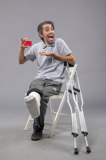 Vue de face jeune homme avec pied cassé et bandage tenant une carte bancaire sur le mur gris accident masculin torsion douleur au pied humain