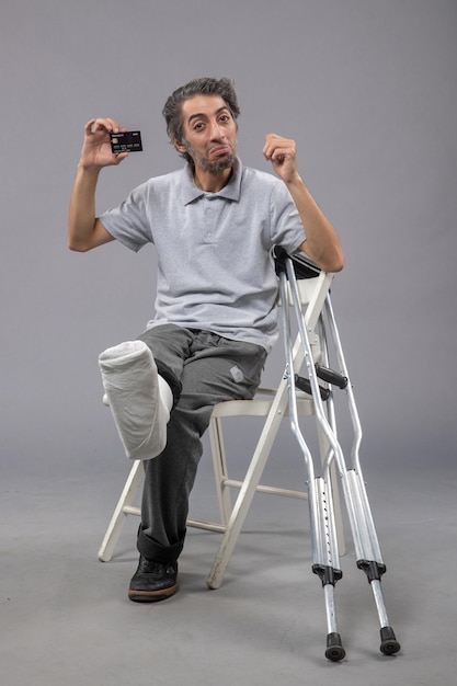 Photo gratuite vue de face jeune homme avec pied cassé et bandage tenant une carte bancaire noire sur le mur gris torsion homme douleur au pied accident