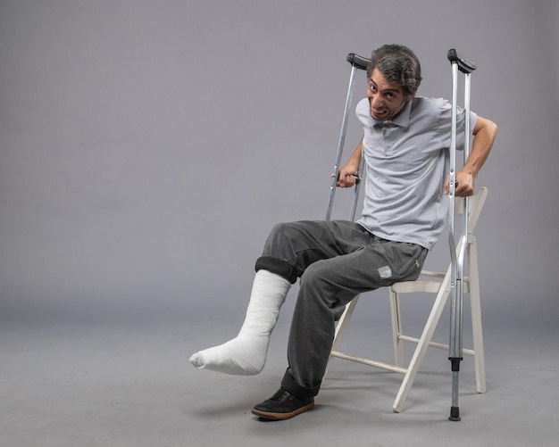 Photo gratuite vue de face jeune homme avec pied cassé essayant de se lever tenant des béquilles sur un bureau gris pied cassé douleur torsion jambe accident