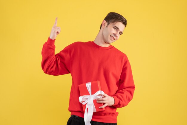 Vue de face jeune homme avec pull rouge pointant avec le doigt vers le haut sur fond jaune