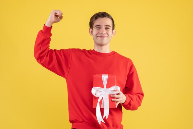 Vue de face jeune homme avec pull rouge tenant un cadeau sur le lieu de copie de fond jaune