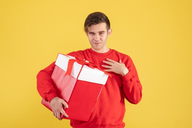 Vue de face jeune homme avec pull rouge tenant un cadeau de Noël sur fond jaune