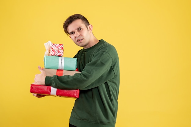 Vue de face jeune homme avec pull vert cachant ses cadeaux debout sur jaune