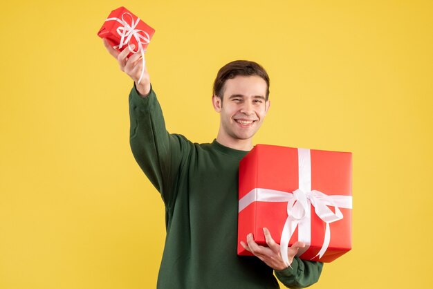 Vue de face jeune homme avec pull vert tenant de grands et petits cadeaux debout sur jaune