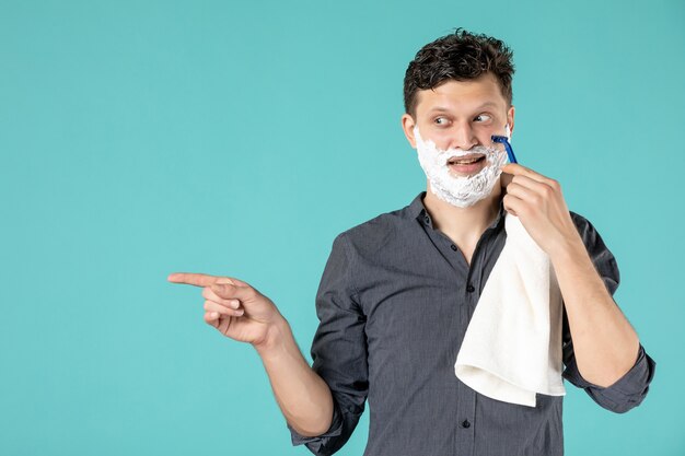 Vue de face jeune homme rasant son visage en mousse avec un rasoir sur fond bleu