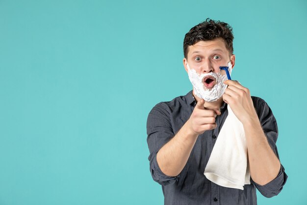 Vue de face jeune homme rasant son visage en mousse avec un rasoir sur fond bleu