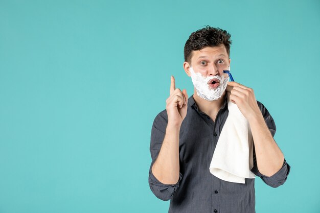 Vue de face jeune homme rasant son visage en mousse avec un rasoir sur fond bleu