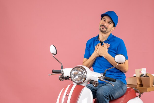 Vue de face d'un jeune homme reconnaissant un coursier portant un chapeau assis sur un scooter en gardant les mains sur la poitrine sur fond de pêche pastel