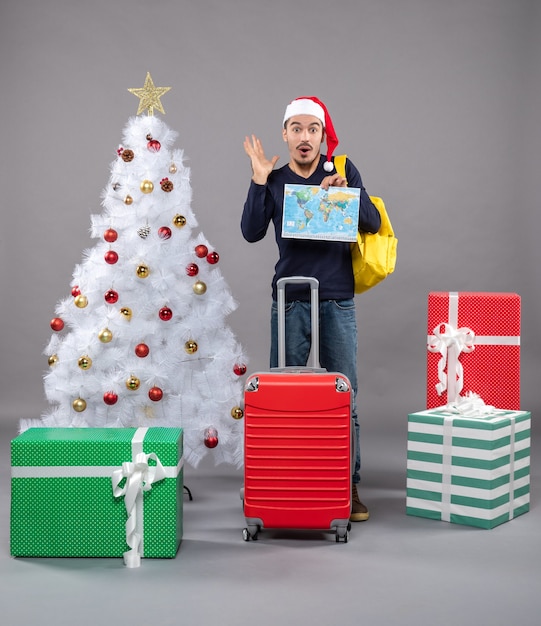 Vue de face jeune homme avec sac à dos jaune tenant la carte près de l'arbre de Noël et présente sur gris isolé