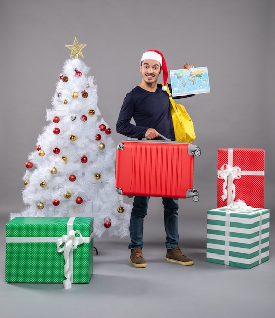 Vue de face jeune homme avec sac à dos jaune tenant une carte et une valise rouge près de l'arbre de Noël et présente sur gris isolé