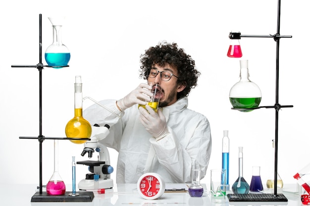 Vue de face jeune homme scientifique en costume spécial assis avec des solutions de boire sur un mur blanc